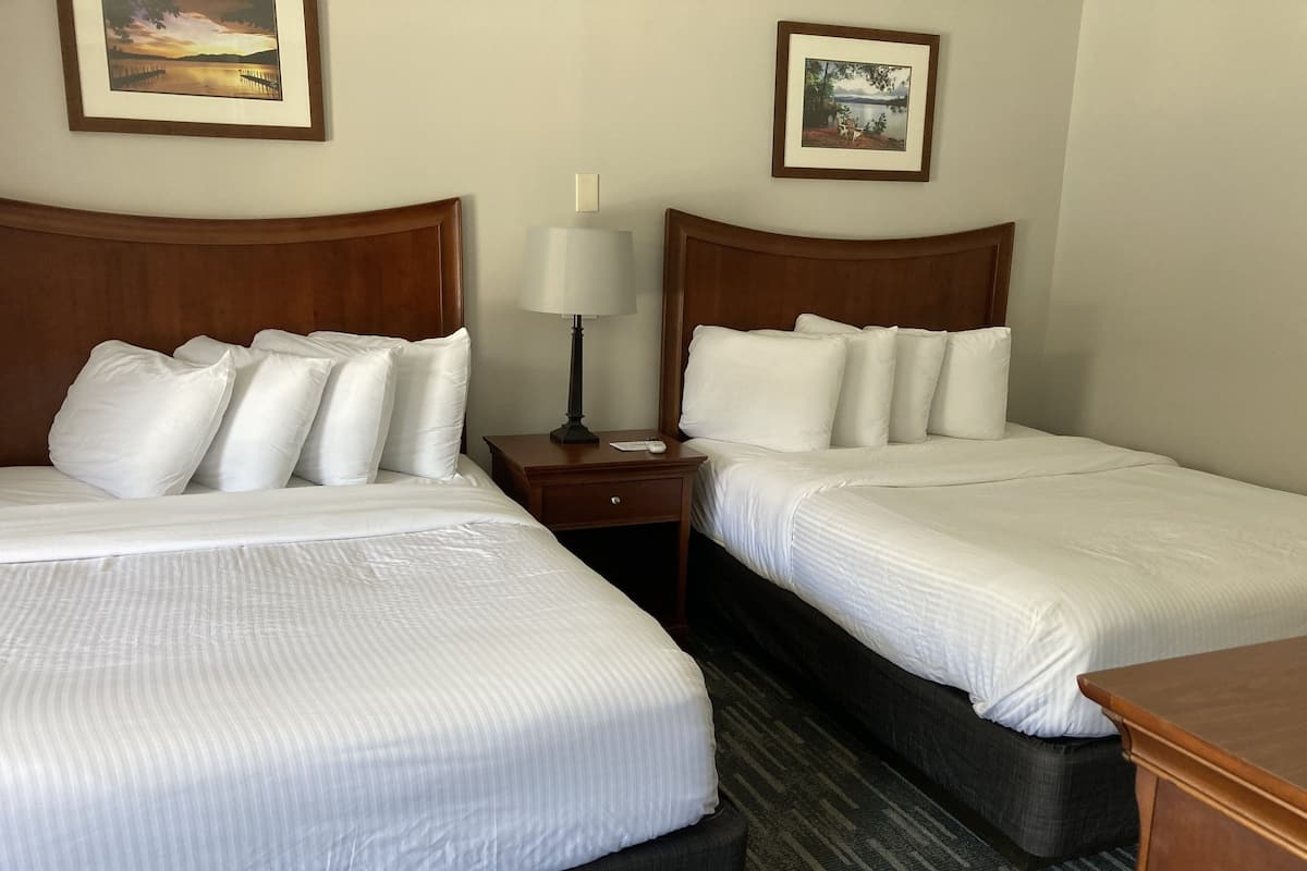 Habitación de hotel con dos camas, cada una con sábanas blancas y varias almohadas. Se observa un mesita de noche con una lámpara y un cuadro enmarcado en la pared sobre cada cama.