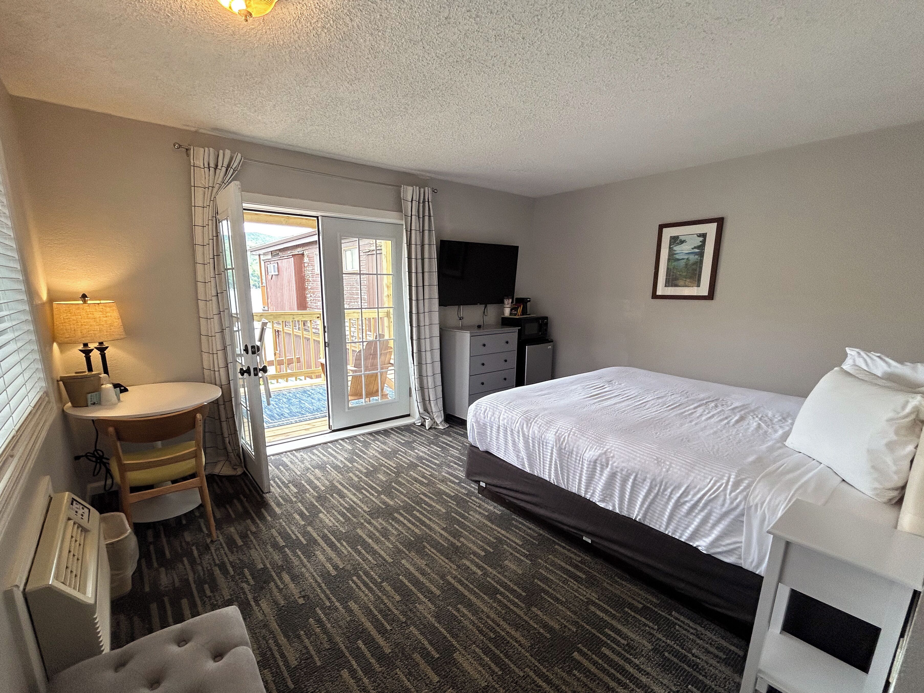 A hotel room with a bed, a desk with a lamp, a chair, a television, and a sliding door to the outside.