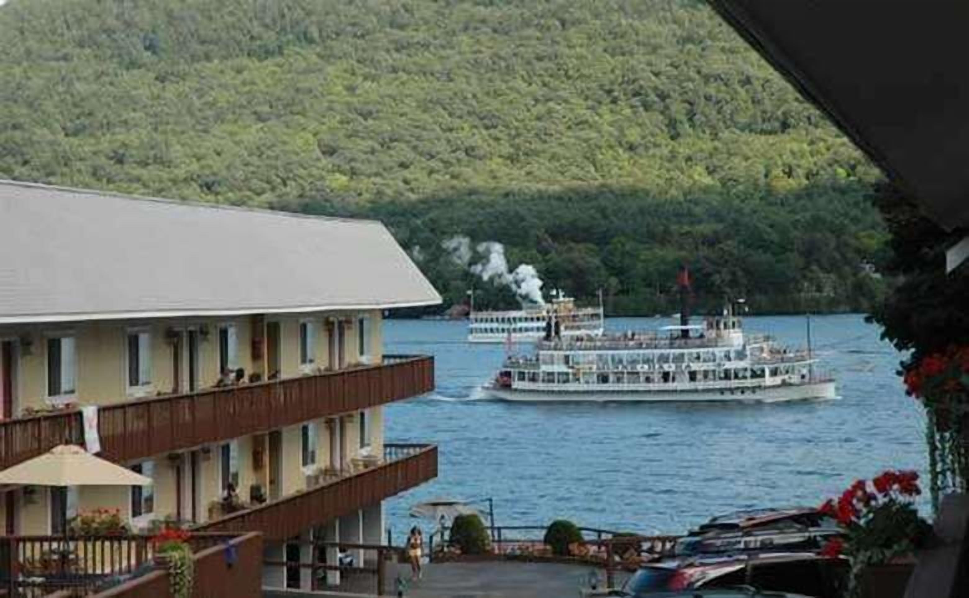 Hotel met balkon met uitzicht op een meer, een boot afgemeerd in de buurt en een beboste heuvel in de achtergrond.