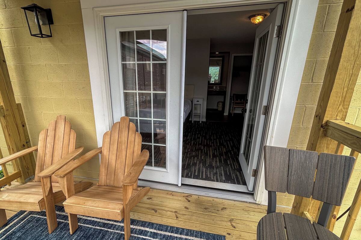 Un porche de madera con dos sillas Adirondack, un panel corredizo que da a una habitación con alfombra estampada y una pequeña mesa blanca.