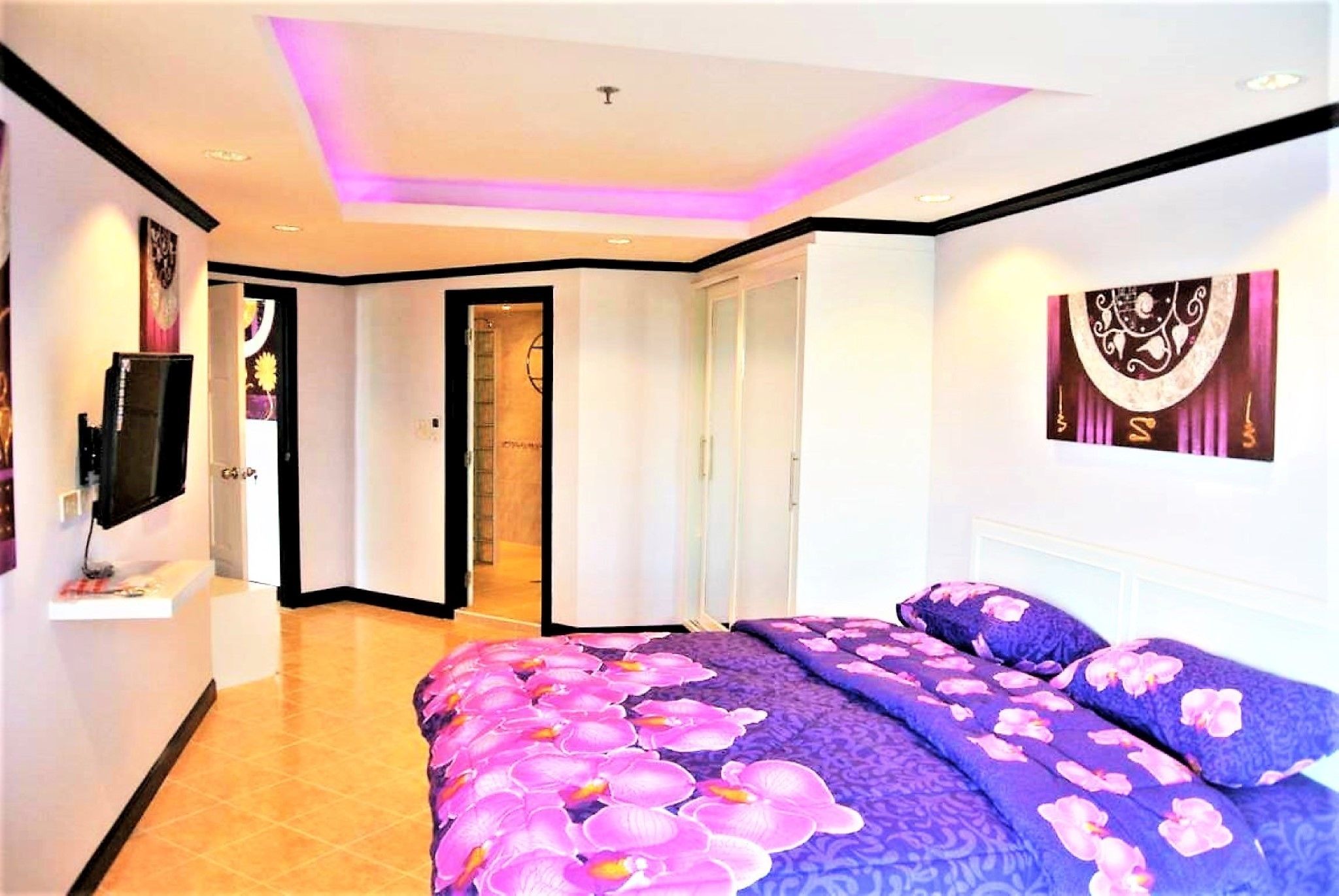 A bedroom with a bed featuring a purple and pink floral bedspread, a flat-screen TV on a stand, and a door leading to a bathroom.