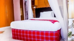 A bed with a white mattress and red plaid skirt, a wooden wardrobe, and a white curtain. - Amboseli Getaway Hotel (Kimana)