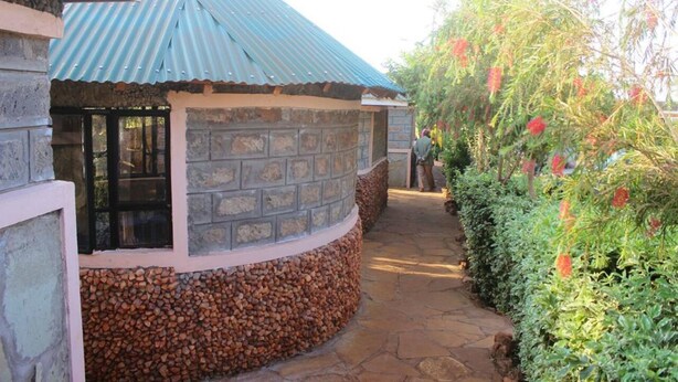 A stone-walled building with a corrugated metal roof, a stone pathway, and a bush with red flowers. - Amboseli Getaway Hotel (Kimana)