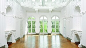 Una habitación amplia con paredes blancas ornamentadas, ventanas arqueadas grandes y un suelo de madera pulida.