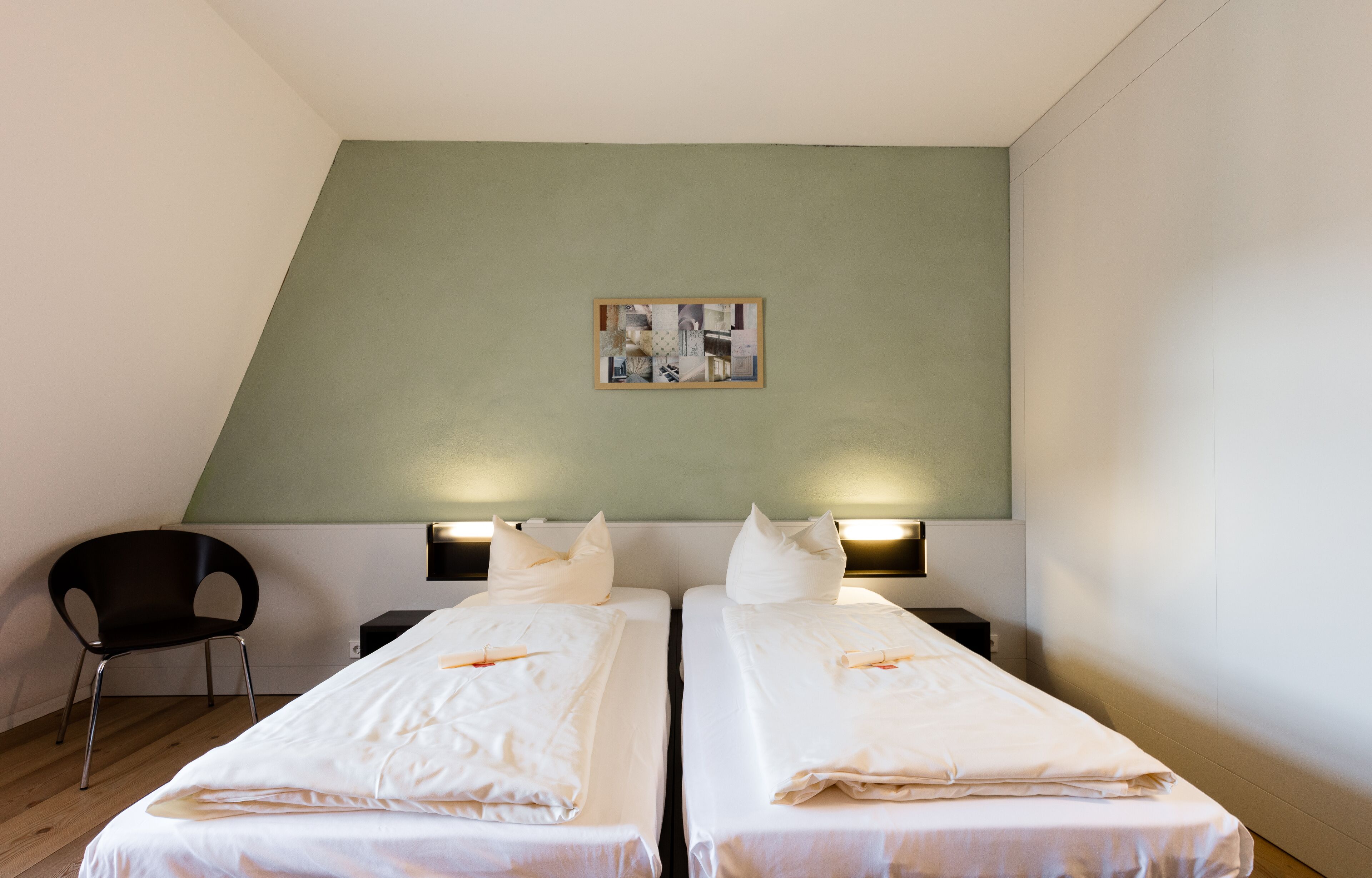 Een hotelkamer met twee enkele bedden, een zwarte stoel, een kleine tafel en een aan de muur hangend schilderij.