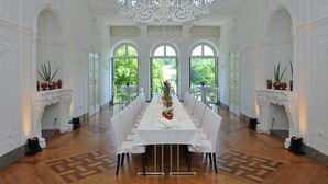 Una larga mesa de comedor con manteles blancos y sillas, ubicada en una habitación con paredes blancas ornamentadas, ventanas amplias y un candelabro.
