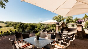 Un patio al aire libre con sillas y mesas de mimbre, un paraguas grande y vistas a un paisaje verde.