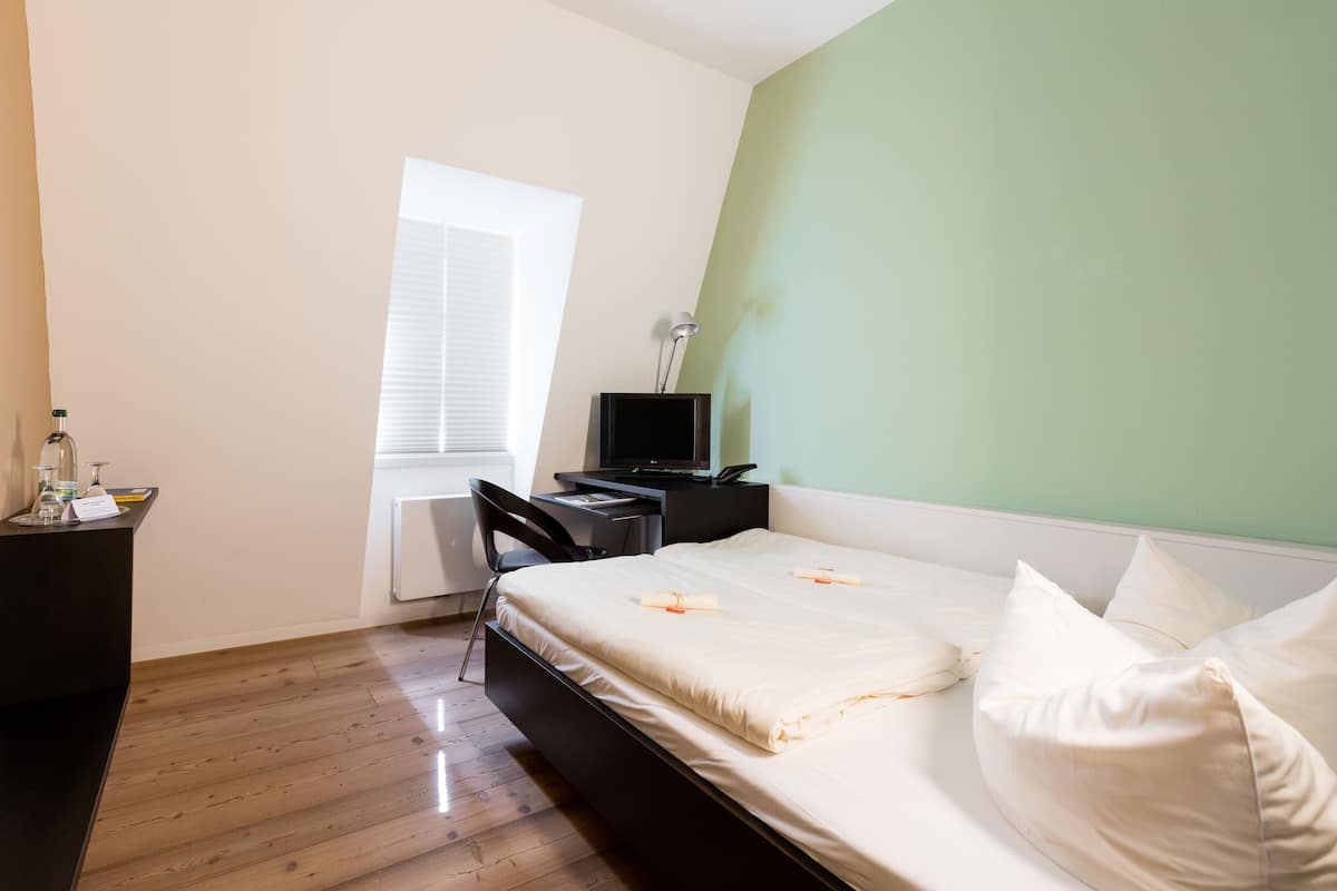 A bedroom with a bed, a desk, a chair, a TV, and a window.
