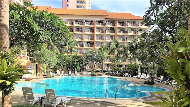 Ein Swimmingpool, umgeben von Liegestühlen und Palmen, mit einem mehrstöckigen Gebäude im Hintergrund.