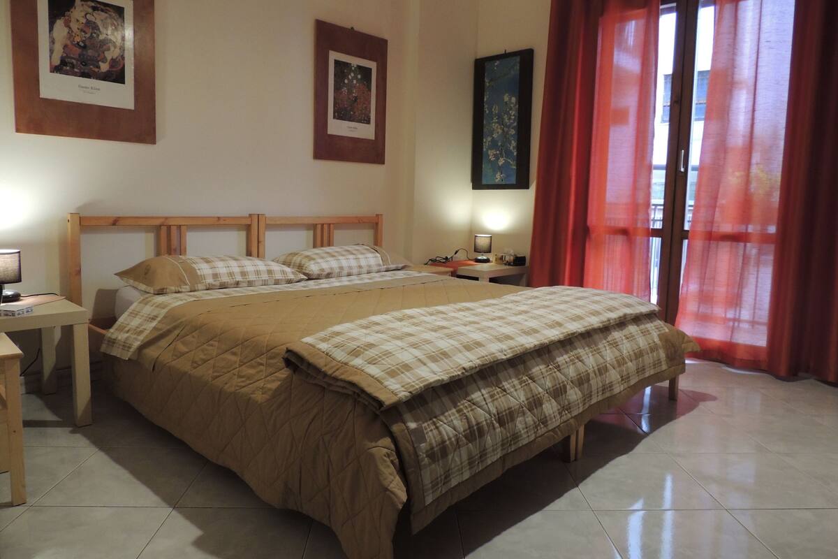 A bedroom with a double bed, a desk with a lamp, and two framed pictures on the wall.