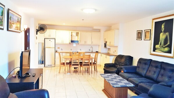 A modern apartment with a living room, dining area, and kitchen. There is a black leather sofa, a dining table with chairs, and a refrigerator.