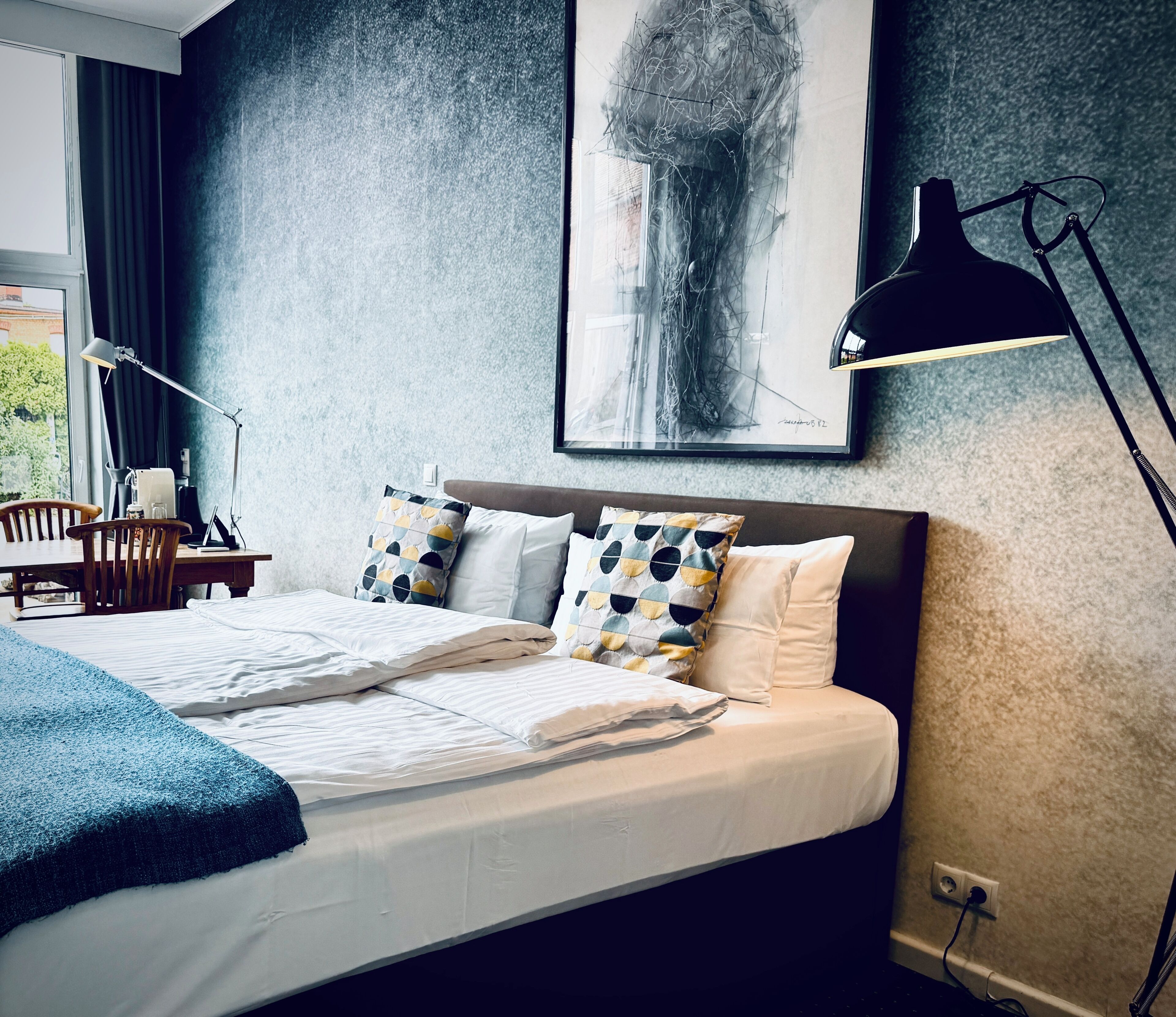 A neatly made bed with white linens, a blue throw blanket, and patterned pillows. A desk with a lamp and a chair is visible to the left.