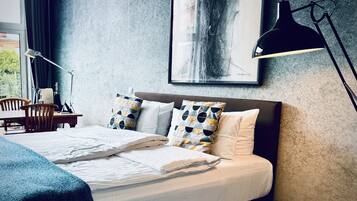 A neatly made bed with white linens, a blue throw blanket, and patterned pillows. A desk with a lamp and a chair is visible to the left.