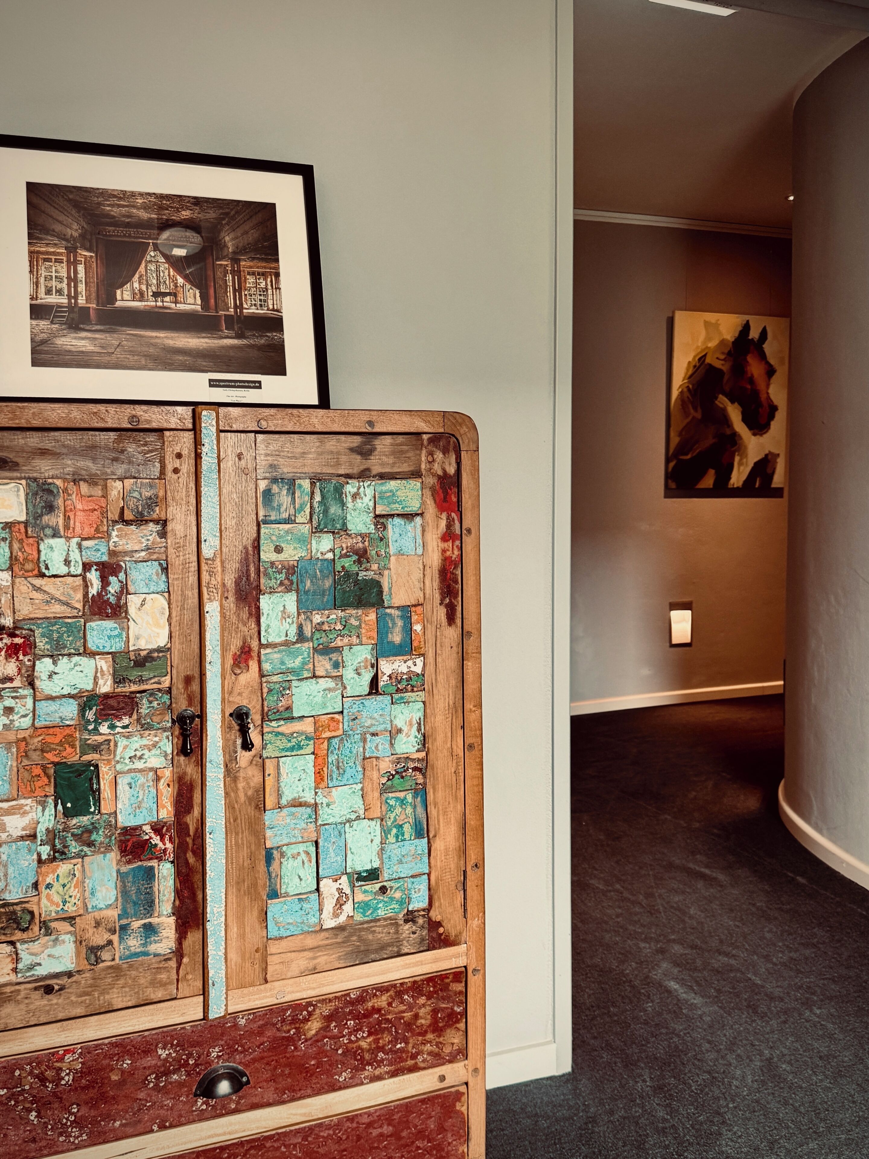 A hallway with a rustic wooden cabinet with a colorful mosaic top, a framed picture on the wall, and a painting on the adjacent wall.