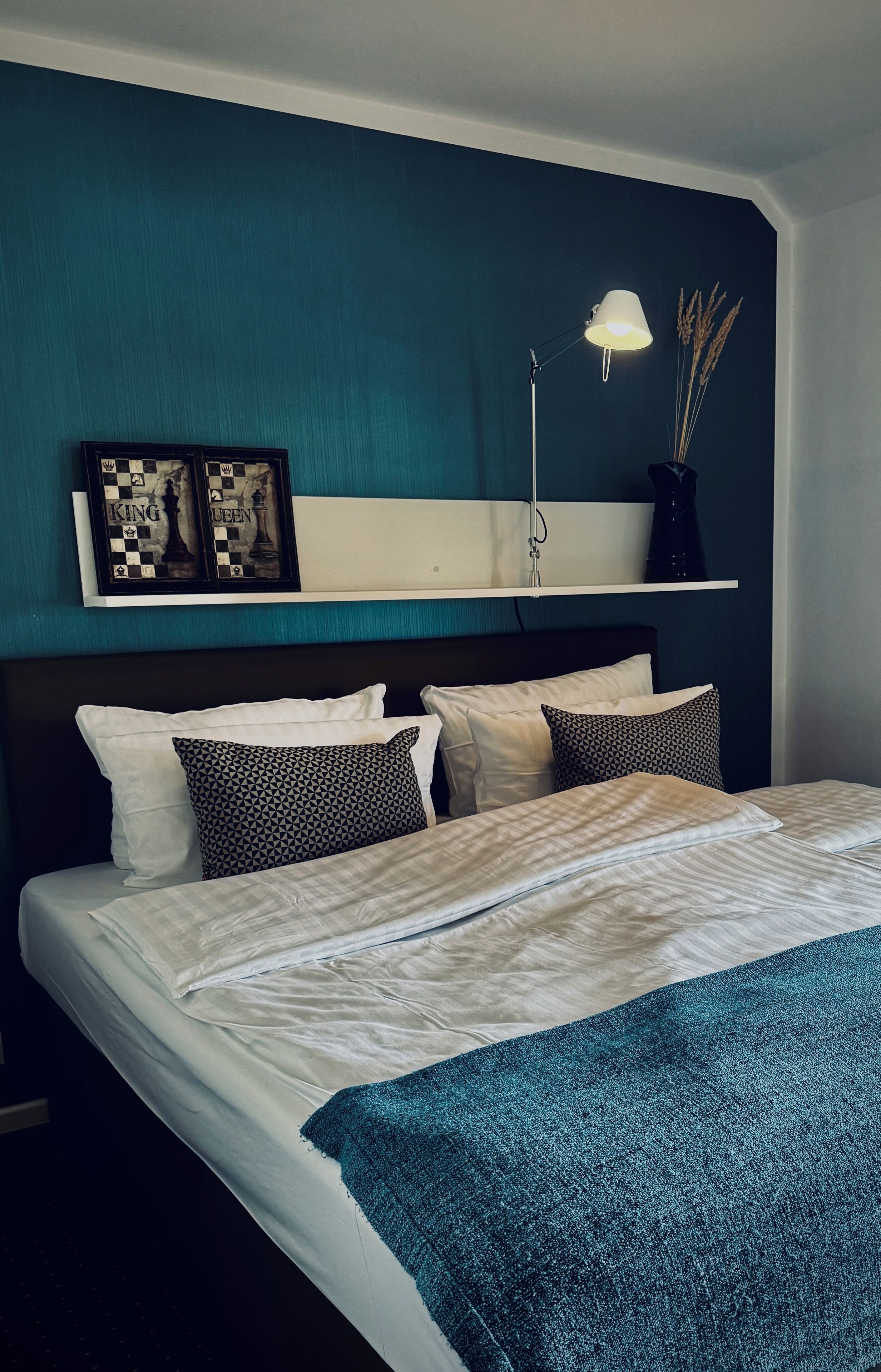 Ein ordentlich bezogenes Bett mit blauer Decke, einem Kopfteil und einem Wandregal mit einem Schachbrettbild und einer Lampe.
