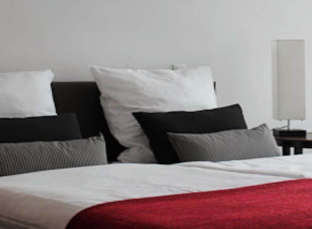 A bed with white and red bedding, a black headboard, and a nightstand with a lamp.