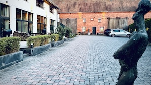 A paved courtyard with a statue, a building with large windows, and a parked car. - Aparthotel Flughafen Leipzig (Schkeuditz)