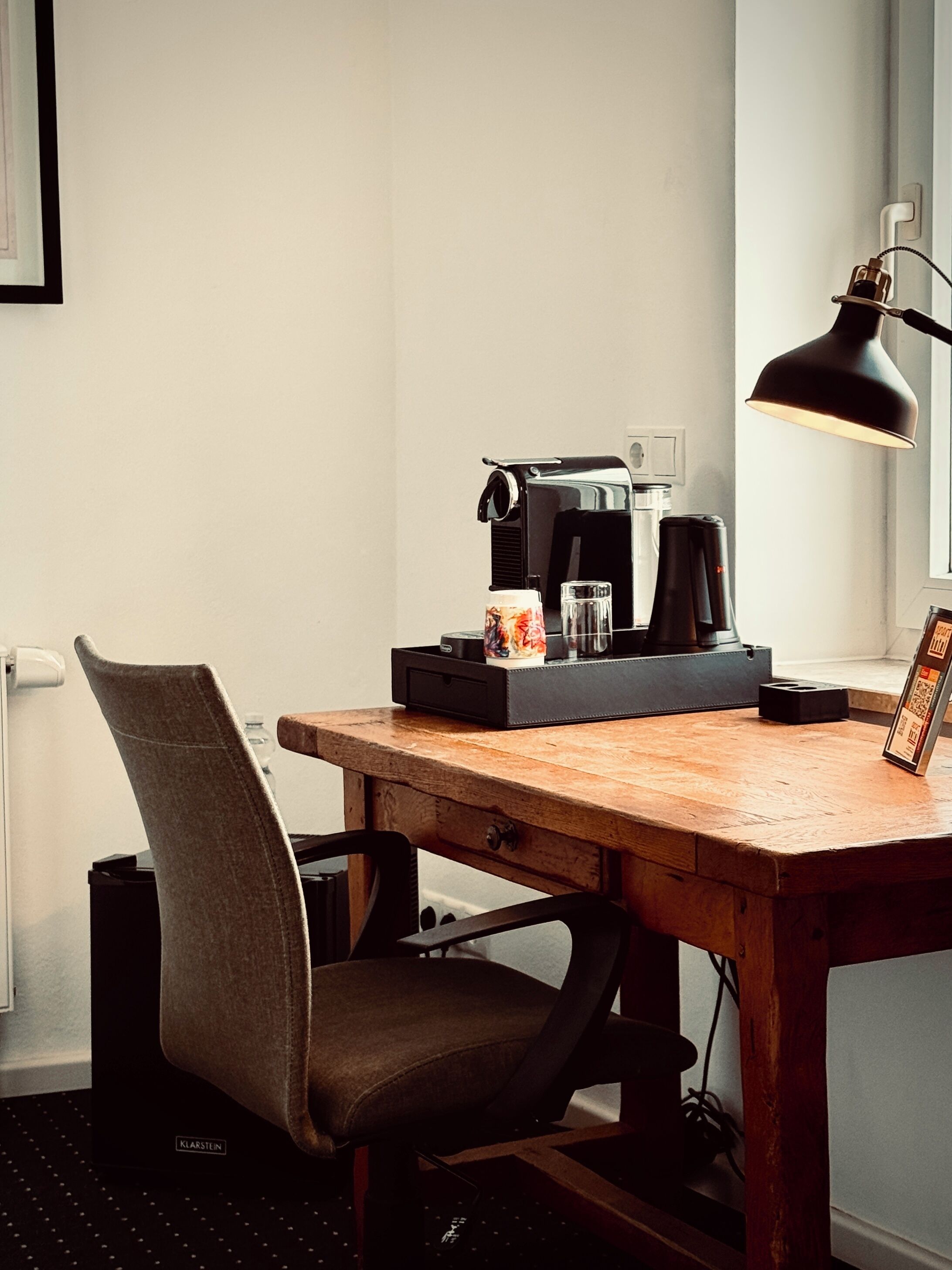 Een houten bureau met een koffiezetapparaat, mok en lamp. Er staat een stoel met een kussen bij.