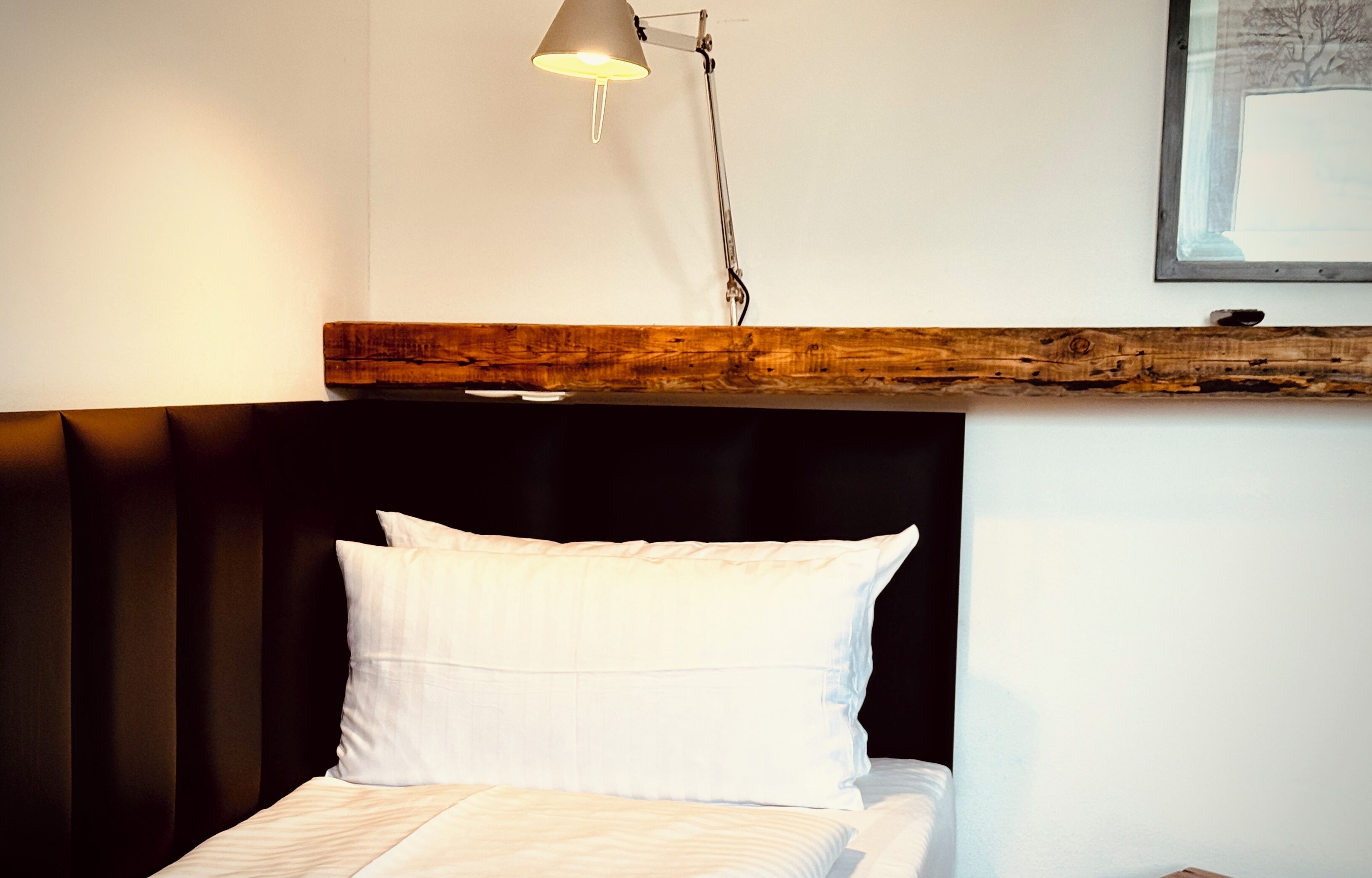 Cama com roupa de cama branca, cabeceira preta e prateleira de madeira acima.
