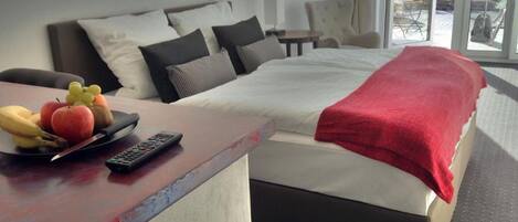 Habitación de hotel con una cama, una mesita de noche con fruta y un control remoto, y vistas al exterior a través de una ventana.