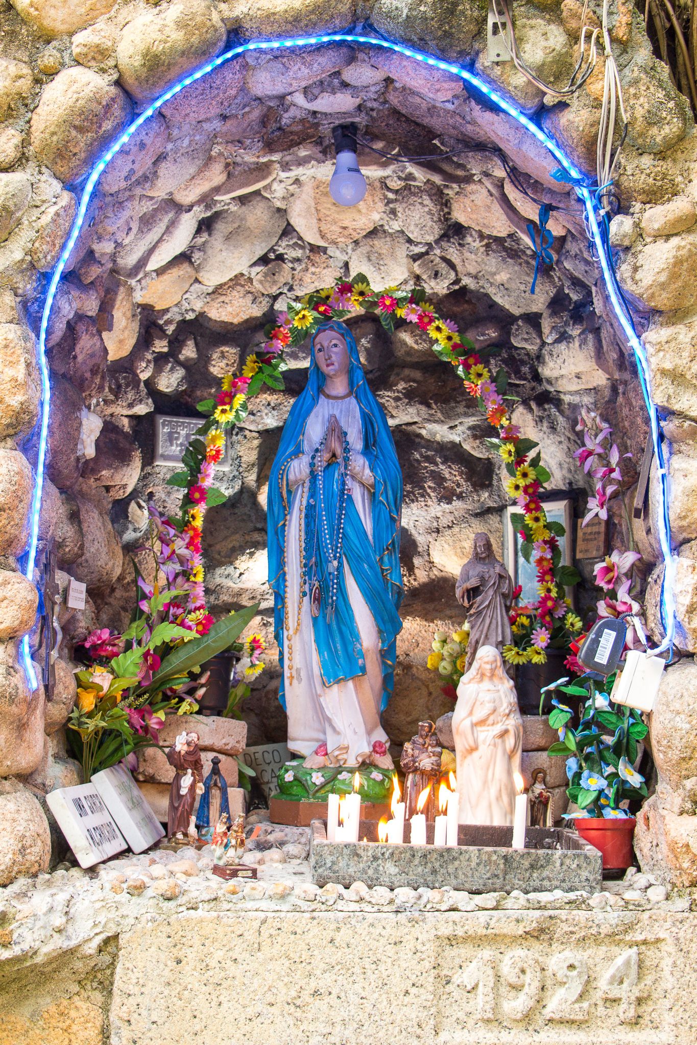 Santuario con estatua, velas, flores y una placa con fecha.