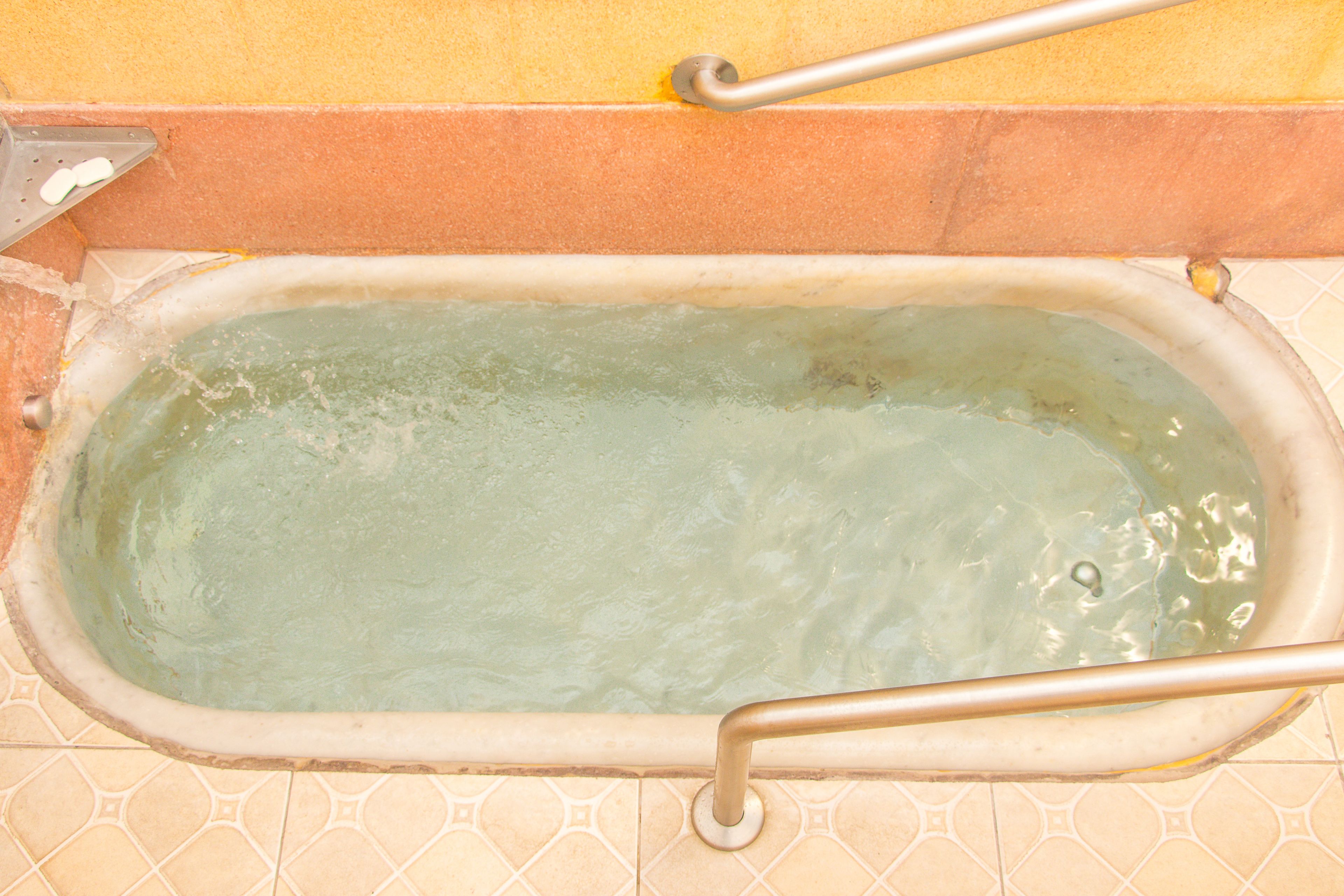 A bathtub with a metal grab bar, tiled walls, and a yellowish-brown floor.