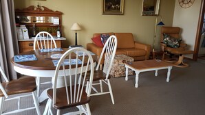 A living room with a dining table, chairs, a sofa, a coffee table, and a side table. There are framed pictures on the walls and a clock.