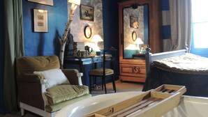 Une pièce avec un mur bleu, des poutres en bois au plafond, un bureau avec une lampe, une chaise, un lit avec une tête de lit en bois et un tapis à motifs.