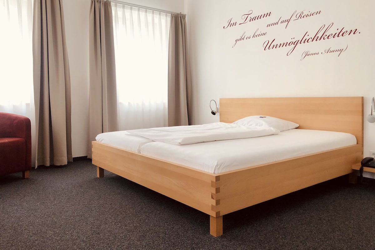 Ein Hotelzimmer mit einem hölzernen Bett, beigen Vorhängen und einem Wandspruch in Deutsch.
