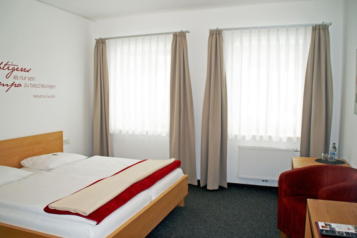 Ein Hotelzimmer mit einem Bett, einem roten Sessel, einem kleinen Tisch mit Fernbedienung und einer Wand mit einem Zitat.