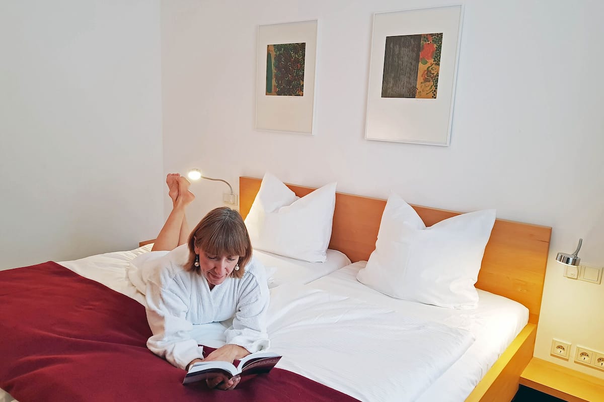 Eine Person liest ein Buch auf einem Bett mit weißer Bettwäsche und einer roten Decke. Über dem Bett befinden sich zwei Wandbilder.