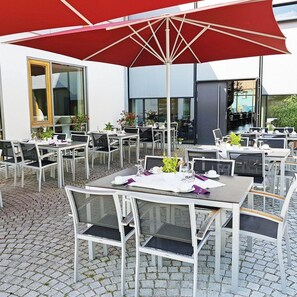 Un área de comedor al aire libre con mesas y sillas, un paraguas rojo y un edificio con grandes ventanales al fondo.