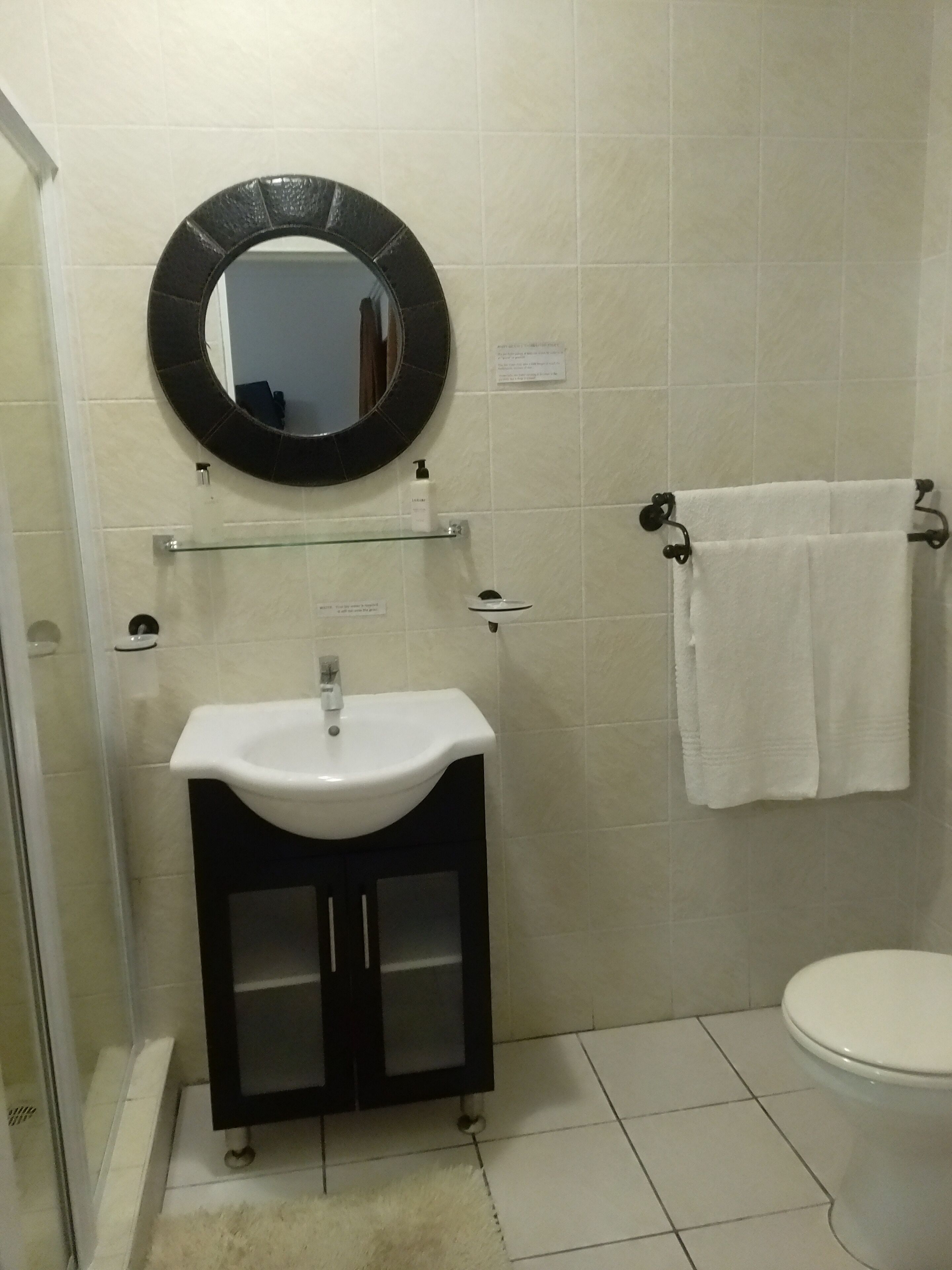 A bathroom with a white sink, a round mirror, a glass shelf, a towel rack, and a toilet.