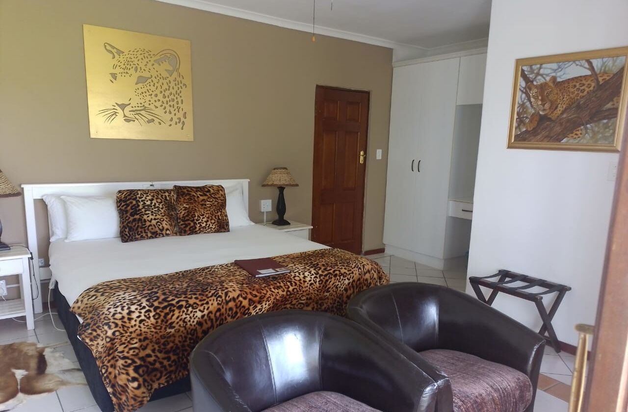 Un dormitorio con una cama, cubrecama estampado de leopardo, dos sillones de cuero, una mesita de noche, una lámpara y dos cuadros con estampado de leopardo en las paredes.