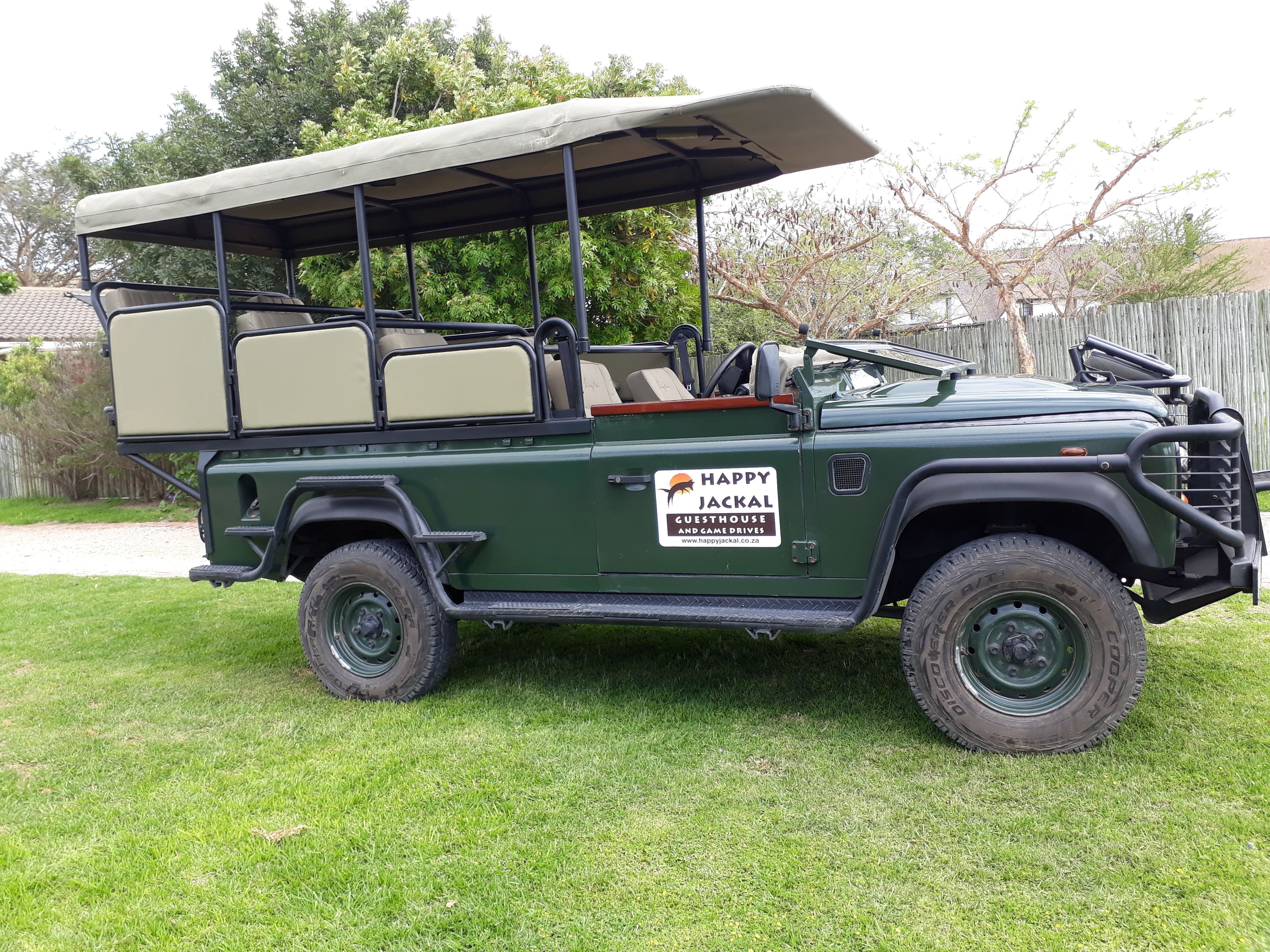 Un vehículo safari verde con toldo, que dice: 'HAPPY JACKAL GUESTHOUSE AND GAME DRIVES'.