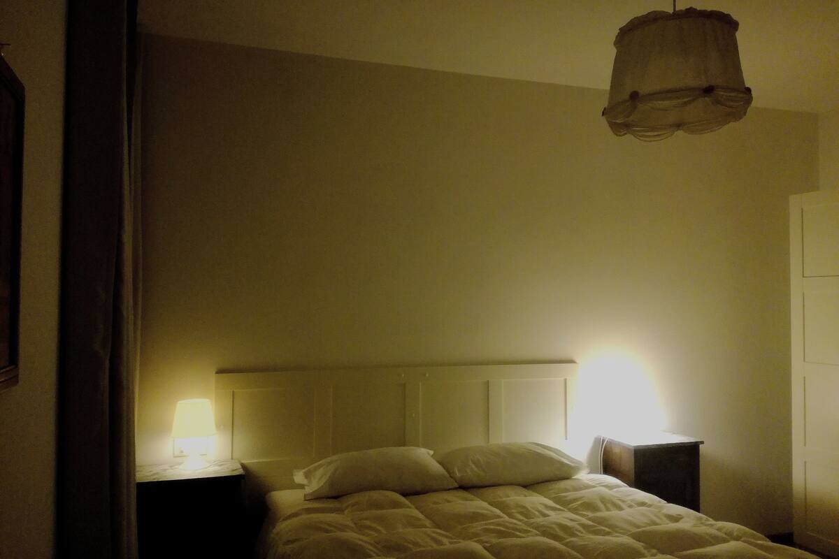 A dimly lit bedroom with a bed, a bedside table, and a hanging light fixture.