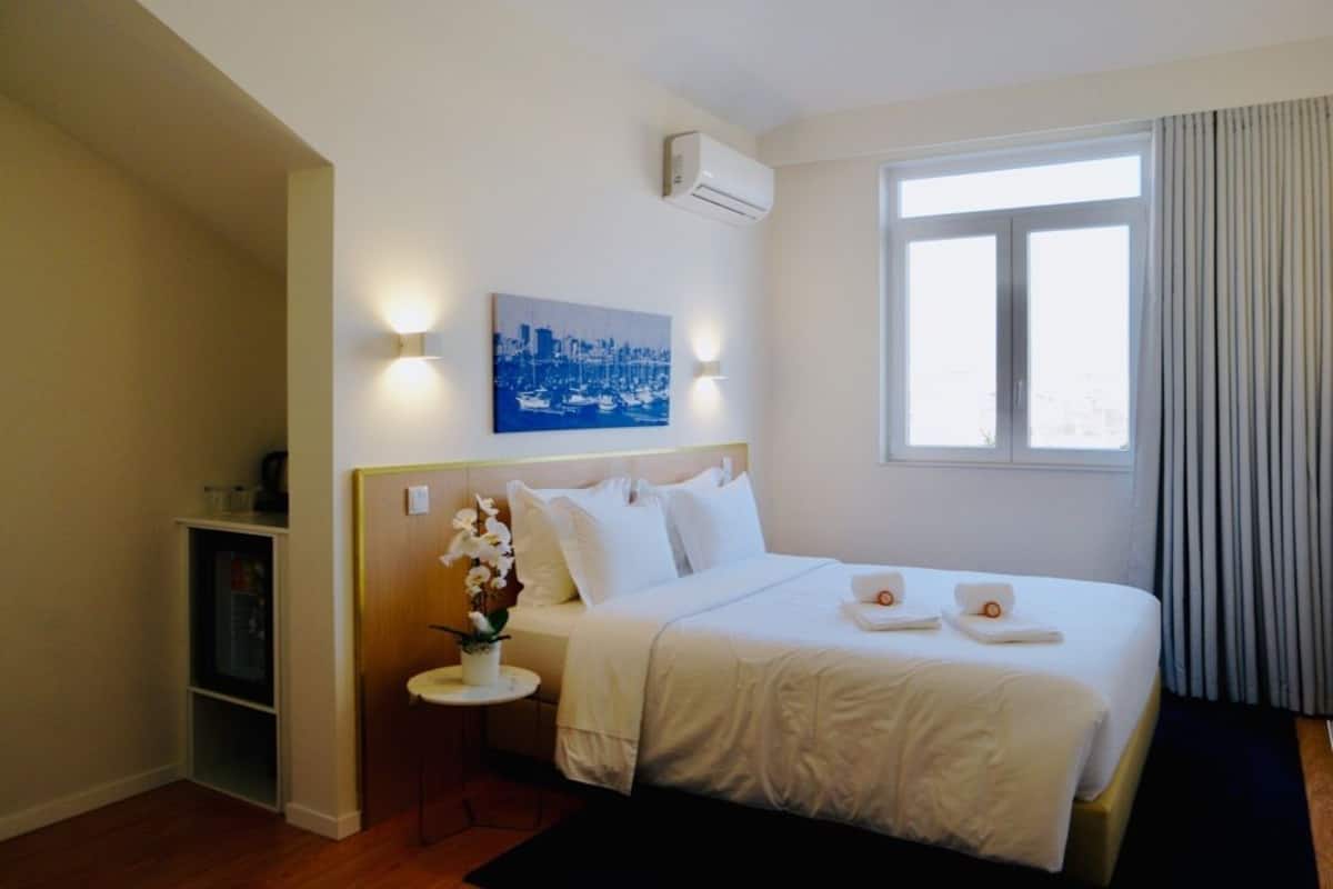 A hotel room with a bed, a small bedside table with a vase of flowers, a wall-mounted air conditioner, a window with curtains, and a cityscape painting above the bed.