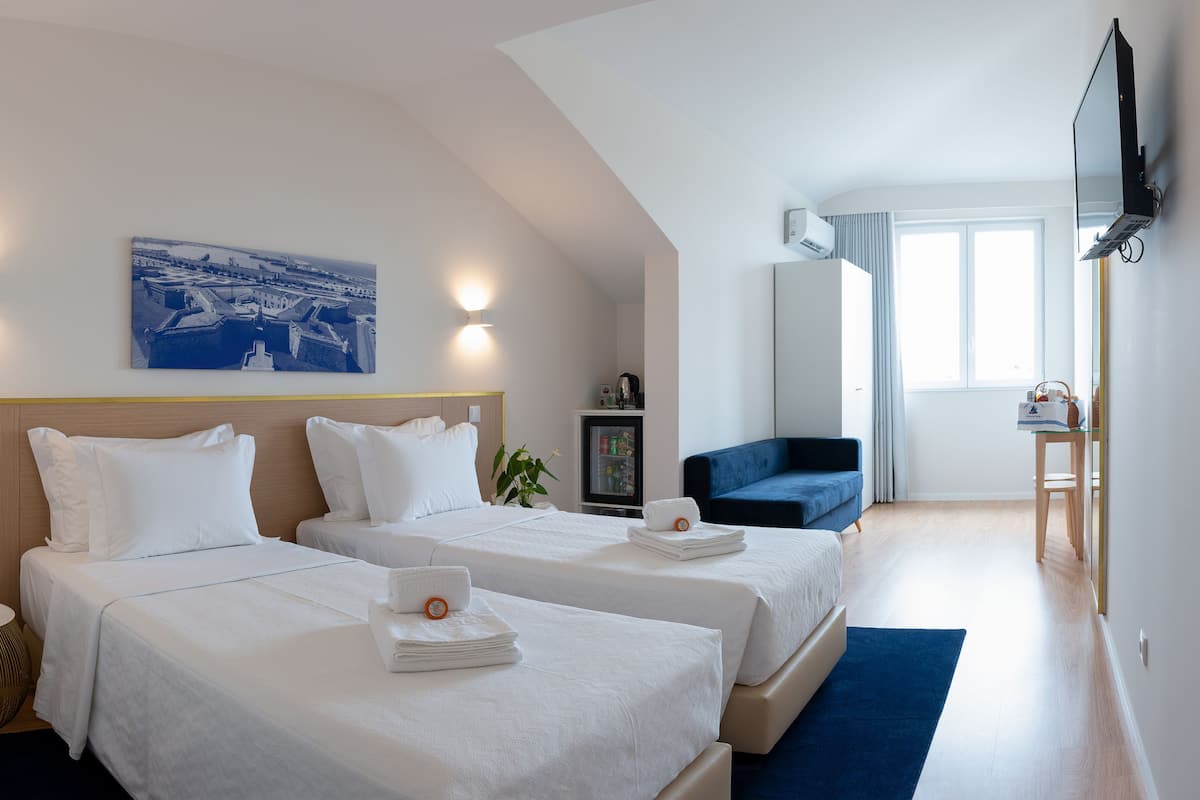 A hotel room with two beds, a small table, a chair, a TV, and a painting on the wall.