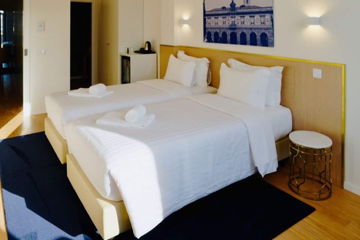A double bed with white linens and towels, a bedside table, a round stool, and a wall-mounted light fixture.