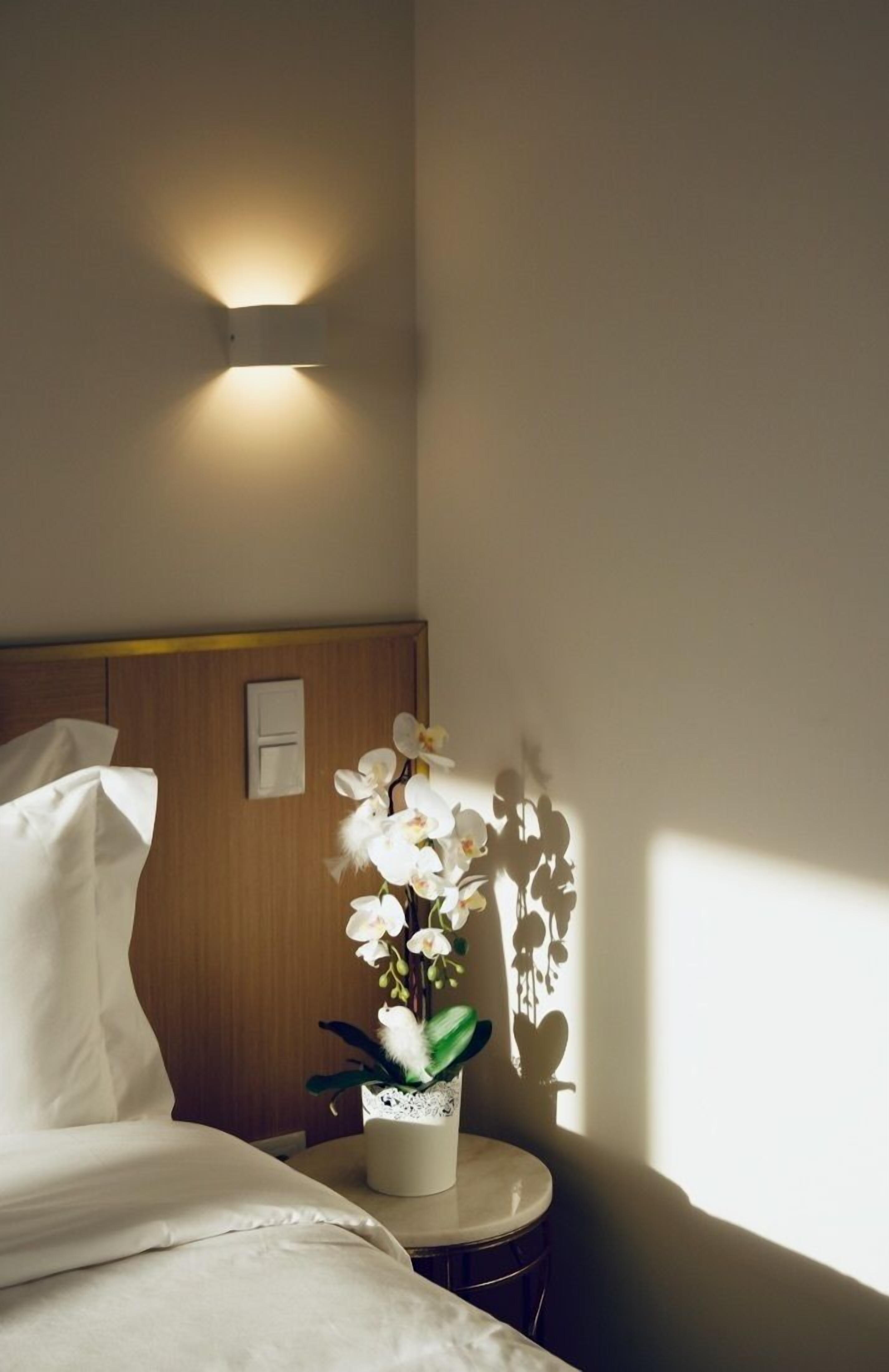 Una habitación de hotel con una cama, una mesita de noche con una orquídea en maceta, una lámpara de pared y un cabecero de madera.