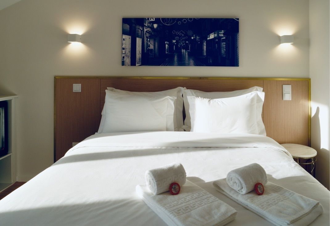 A hotel room with a large bed, two pillows, a headboard, two wall-mounted lights, a painting above the bed, and two rolled towels on the bed.