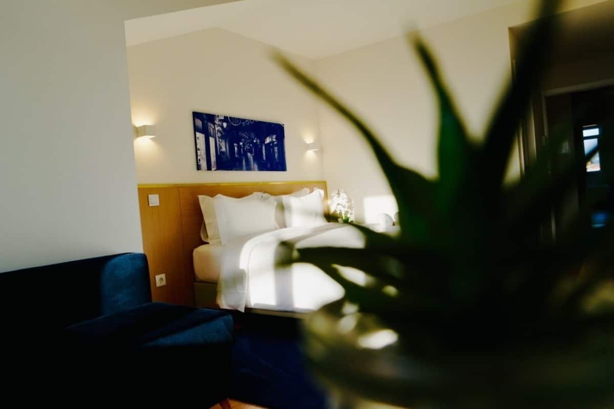 A hotel room with a bed, a blue armchair, a potted plant, and a wall-mounted artwork.