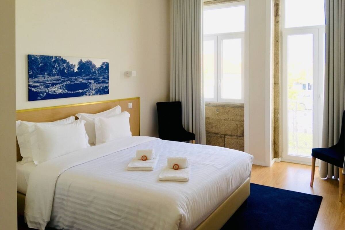 A hotel room with a large bed, a painting on the wall, and a view through the window.