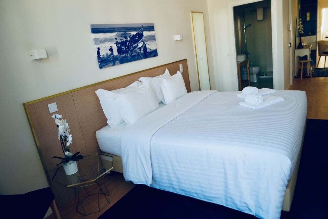 A hotel room with a large bed, a small table with a vase of flowers, a painting on the wall, and a bathroom visible through an open door.