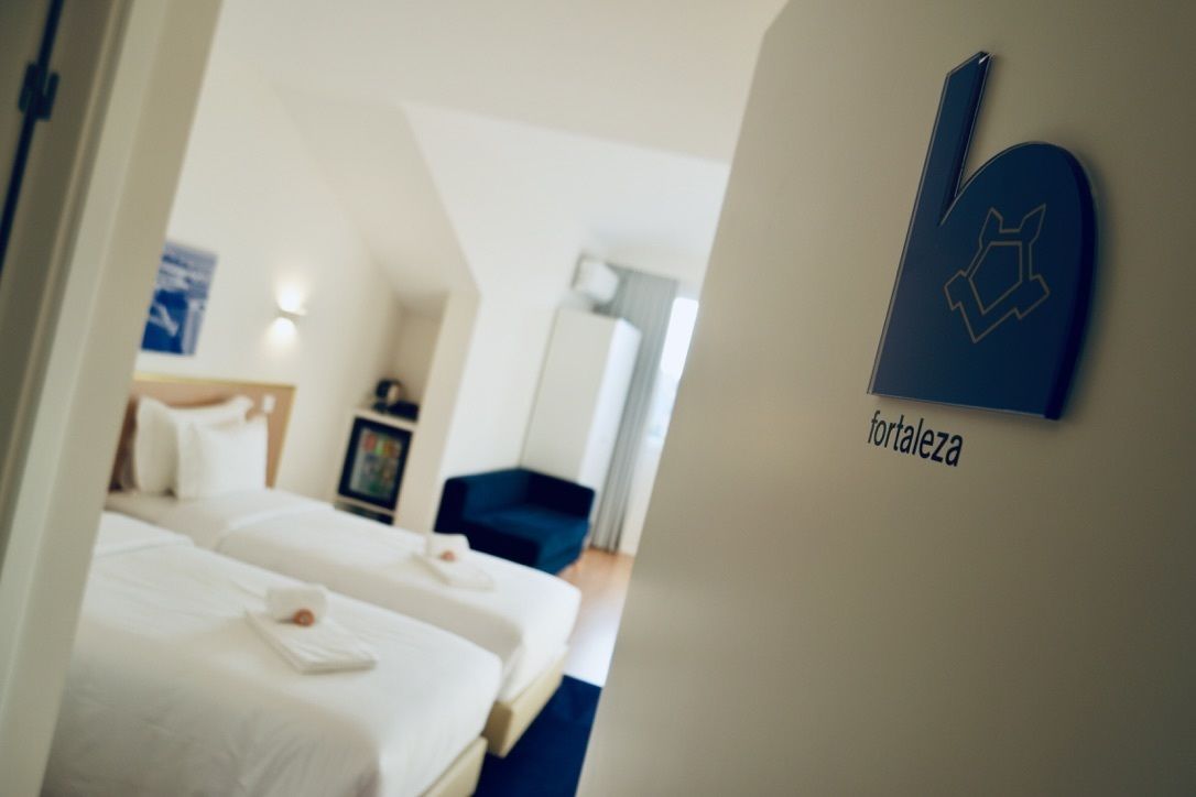 A hotel room with two beds, a blue chair, and a wall-mounted sign reading "fortaleza".