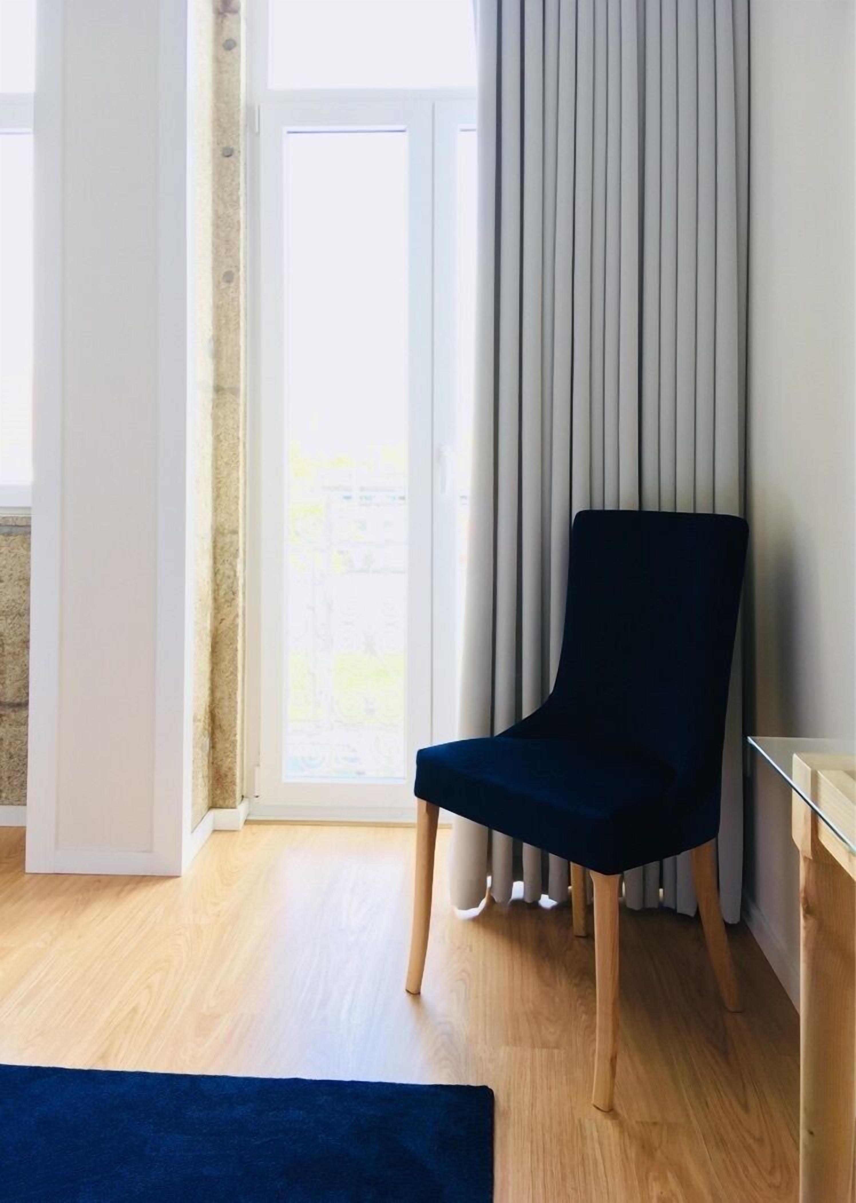 A room with a dark blue chair, a wooden floor, a glass table, and a view through a window with curtains.