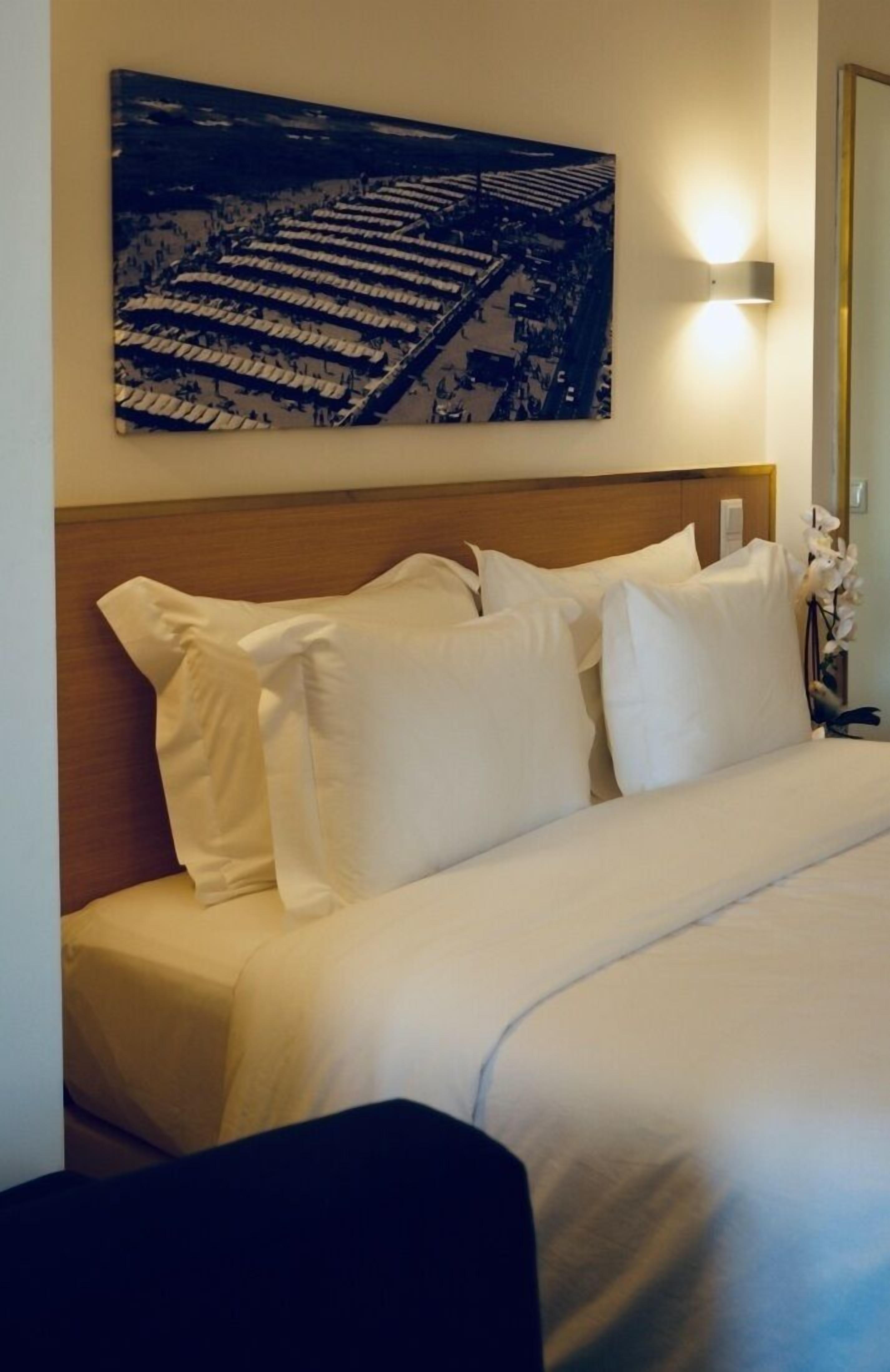 Une chambre d’hôtel avec un lit bien fait, une tête de lit, une lampe fixée au mur et une œuvre d’art encadrée au-dessus du lit.