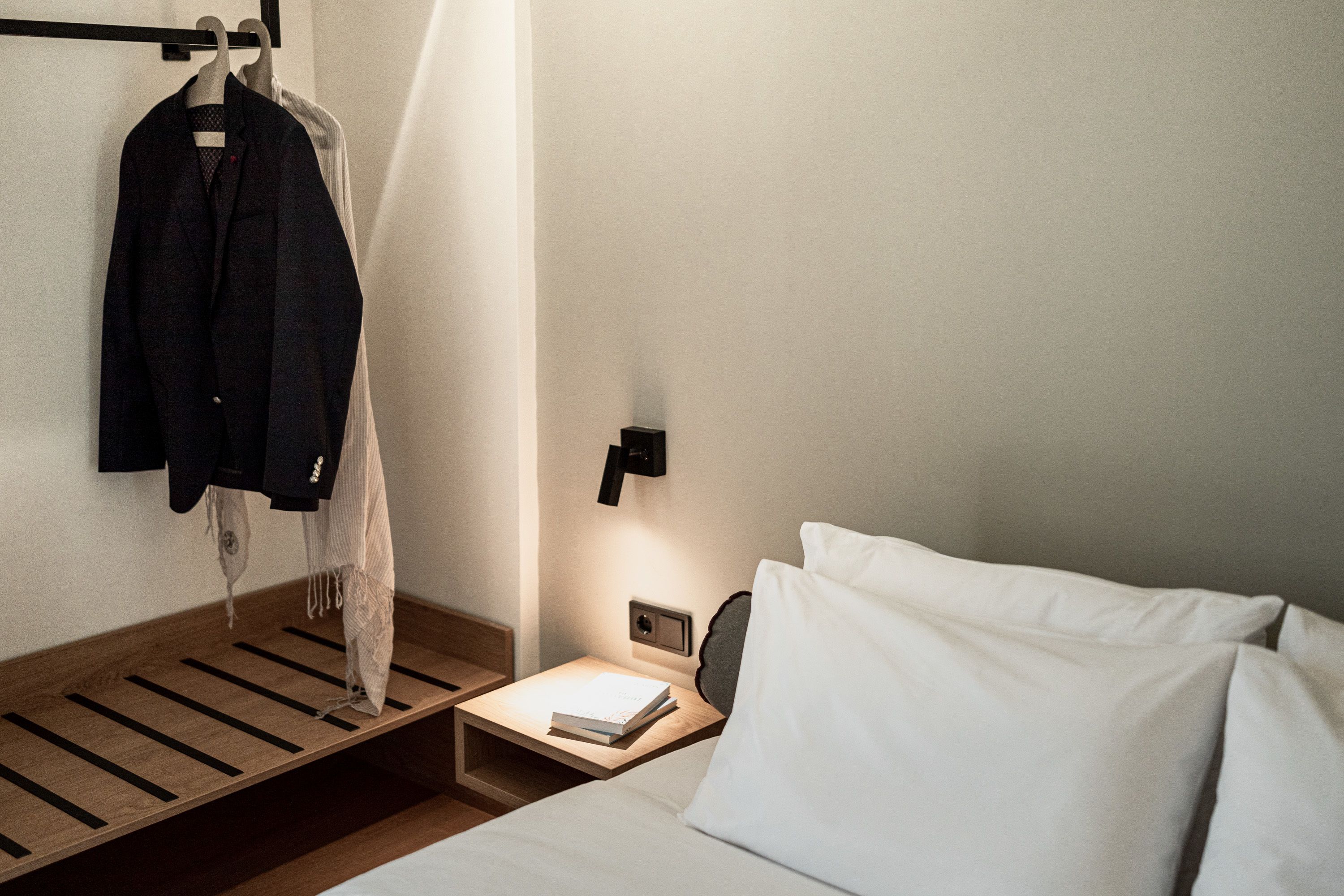 Une chambre d’hôtel bien rangée, avec un lit, une table de chevet sur laquelle repose un livre, un porte-manteau suspendu et une lampe fixée au mur.