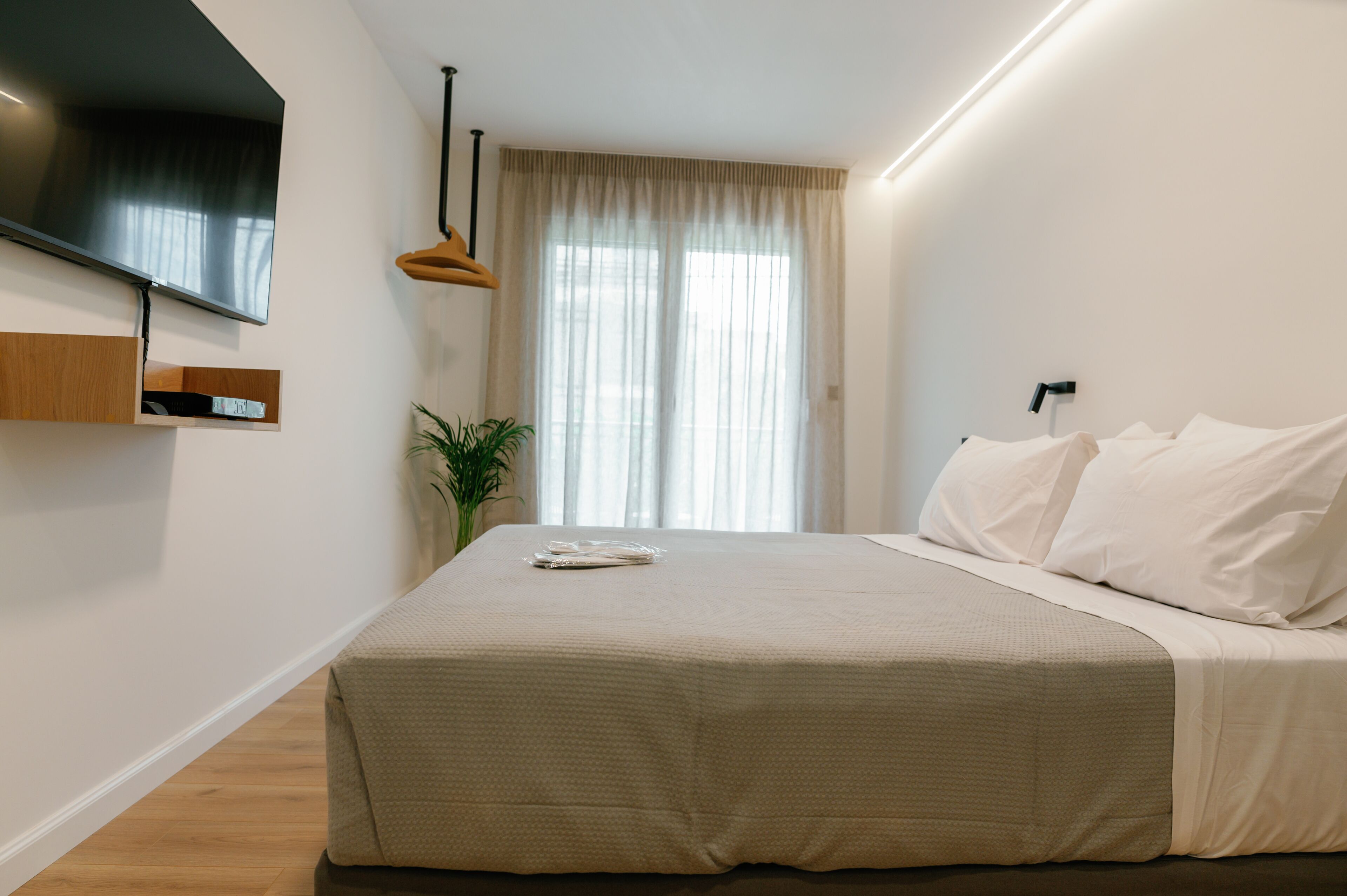 Ein modernes Schlafzimmer mit einem großen Bett, einem Flachbildfernseher an der Wand, einem hölzernen Regal und einer Topfpflanze.