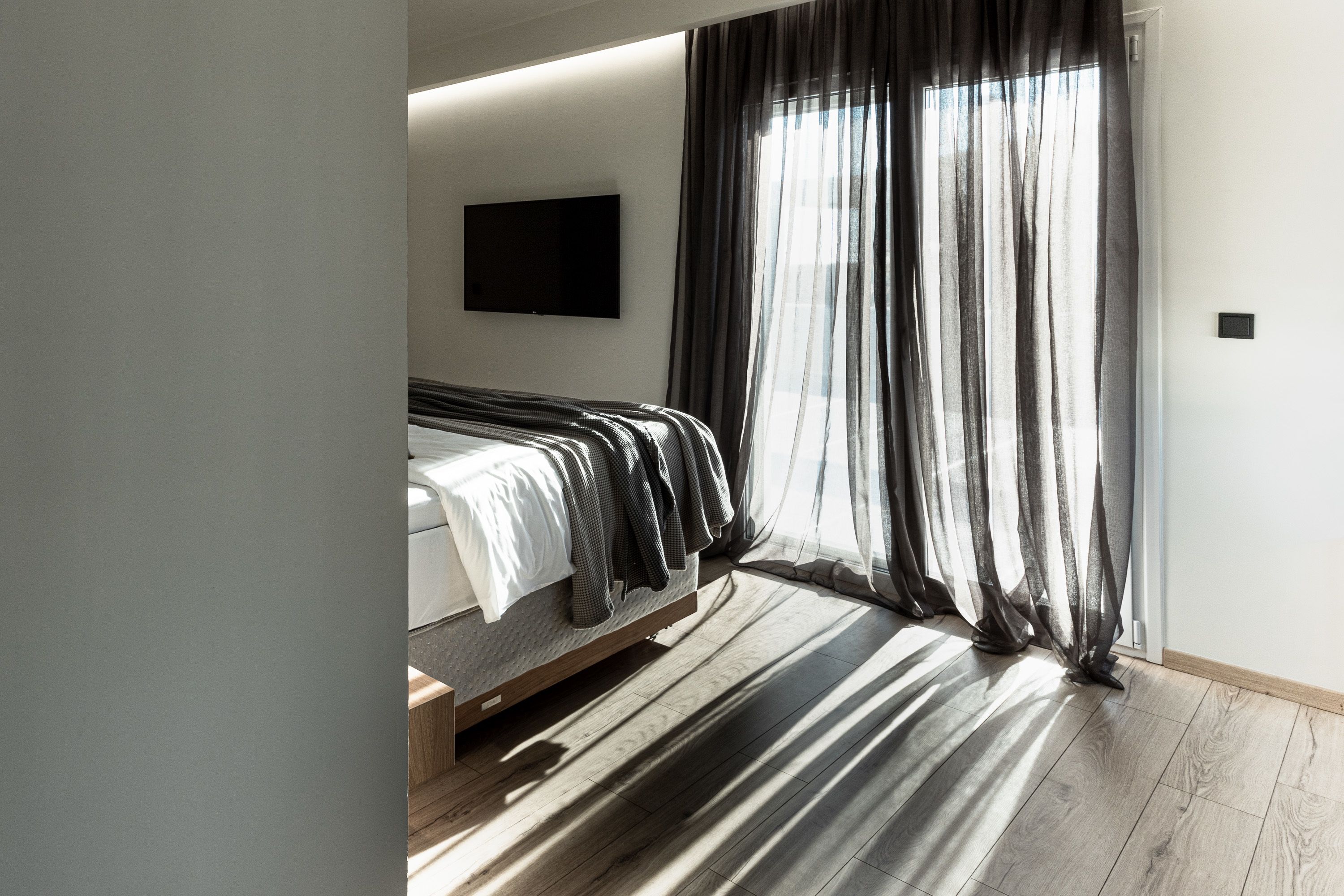 Un dormitorio moderno con una cama, cortinas traslúcidas y un televisor de pantalla plana en la pared.